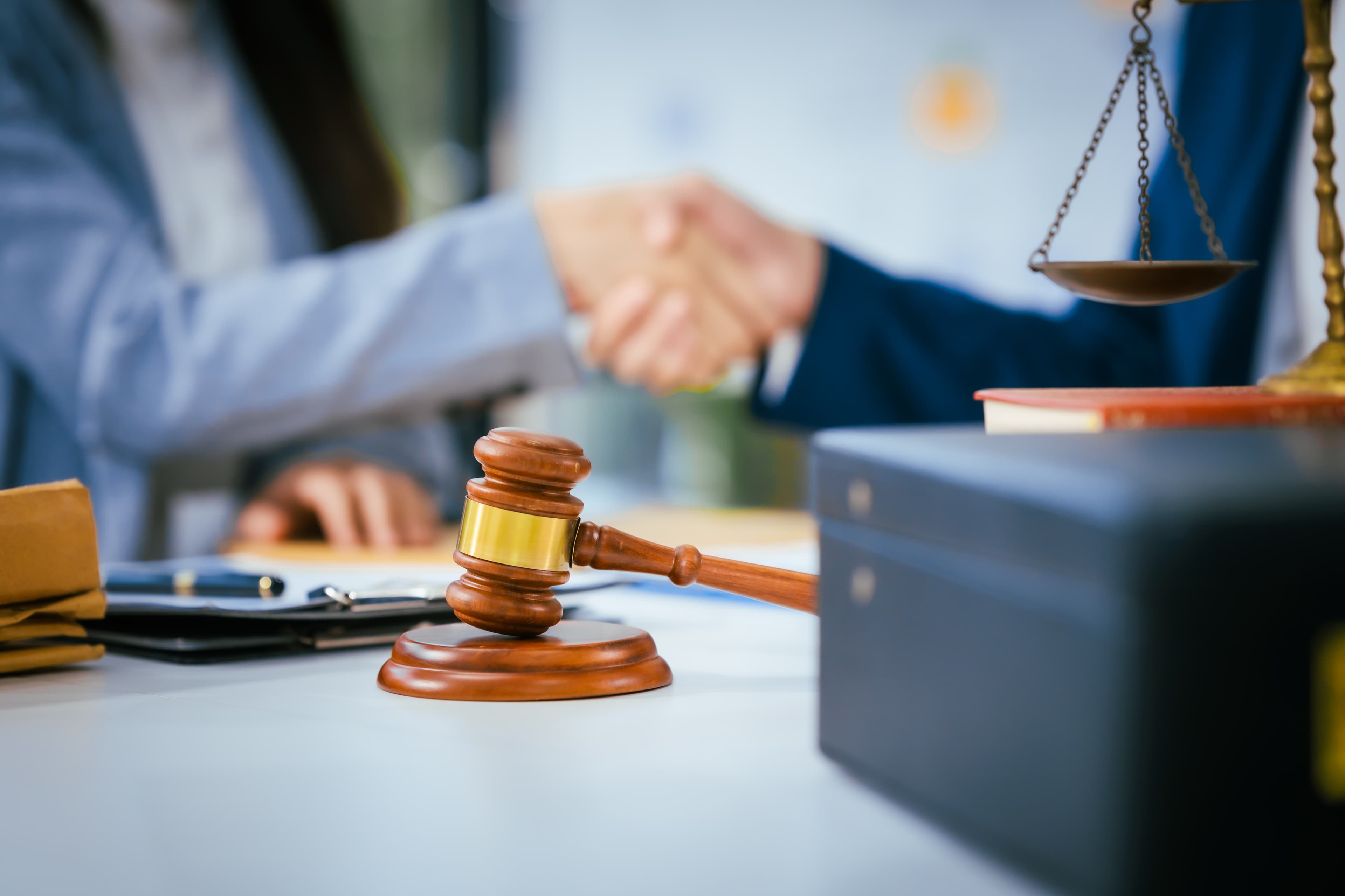 A gavel and scales of justice on a desk with two people shaking hands in the background